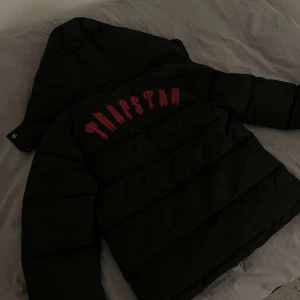Svart pufferjacka från Trapstar - Säljer en svart pufferjacka från Trapstar i mycket bra skick. Jackan har en stor huva och är långärmad med en snygg röd logga på ryggen. Den är perfekt för kalla dagar och har en dragkedja framtill. Passar till alla tillfällen nu när det blir kallt på vintern.