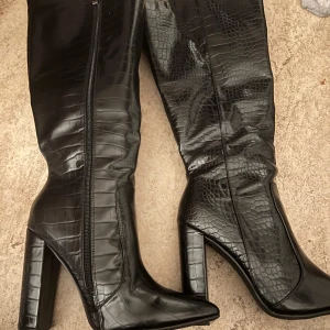Stövlar - Snygga svarta stövlar med krokodil-mönster. Stövlarna har en hög klack och en spetsig tå. De är i gott skick. Köpt för 2000kr använt en gång säljer för 1000