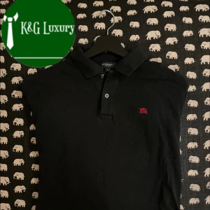 Svart pikétröja från Burberry - Snygg svart pikétröja från Burberry med klassisk krage och knappar. Den har en liten röd broderad logga på bröstet. Passar dig som är 175-185 cm lång! Skicka ett dm om du har några funderingar!