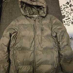 Grön pufferjacka från Kappa - Säljer en grön pufferjacka från Kappa i bra skick. Jackan har en praktisk huva och dragkedja framtill. Perfekt för kyliga dagar och håller dig varm och bekväm. Passar både till vardags och utomhusaktiviteter.