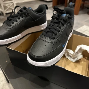 Svarta Nike Air Force sneakers - Säljer ett par svarta Nike Air Force sneakers i nytt skick. De har en klassisk design med vit sula och en liten blå swoosh-detalj. Perfekta för både vardag och träning. Skorna är bekväma och stilrena, passar till det mesta!