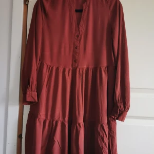 Brun röd klänning från Amisu - Säljer en superfin brun röd klänning från Amisu i storlek XS. Klänningen har långa ärmar och knappar framtill, vilket ger en stilren look. Perfekt för höst och vår! Materialet känns mjukt och bekvämt, perfekt för vardag.