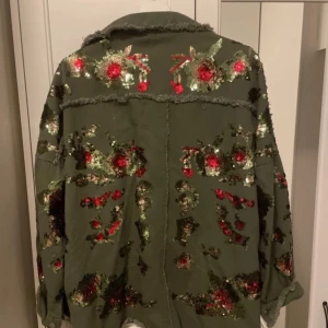 Grön jacka med paljetter - Säljer en unik grön jacka med blommiga paljettmönster i rött och guld. Jackan har en avslappnad passform med fransiga detaljer och knappar framtill. Perfekt för att sticka ut på festen eller till vardags. Storlek är inte angiven, men den ser ut att ha en normal passform.