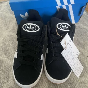 Svarta Adidas Campus sneakers - Säljer ett par snygga svarta Adidas Campus sneakers i nytt skick. De har den klassiska tre-randiga designen i vitt på sidan och en bekväm gummisula. Perfekta för vardagsbruk eller en avslappnad stil. Skorna kommer med extra vita skosnören.