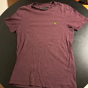 Vinröd t-shirt från Lyle & Scott - Säljer en vinröd t-shirt från Lyle & Scott i bomull. Den har en liten gul logga på bröstet och är kortärmad. Perfekt för vardagsbruk eller en avslappnad dag ute. Skön och stilren design!
