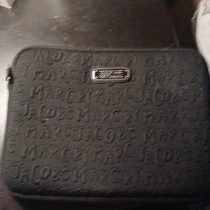 Svart datorväska från Marc Jacobs - Säljer en snygg svart datorväska från Marc Jacobs. Väskan har ett präglat mönster med märkesnamnet och en metallplatta med text. Den är perfekt för att skydda din laptop och har en dragkedja för säker förslutning. Stilren och praktisk för både arbete och skola. Jag säljer den för den är för liten för min dator och jag har köpt den så jag vet att den är äkta. 
