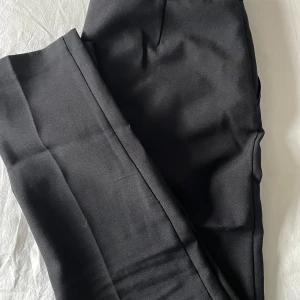 Svarta kostymbyxor - Säljer ett par stilrena svarta kostymbyxor i mycket bra skick. De har en klassisk design med normal passform och är perfekta för både kontoret och festliga tillfällen. Byxorna har en dragkedja och knappar framtill samt hällor för bälte.