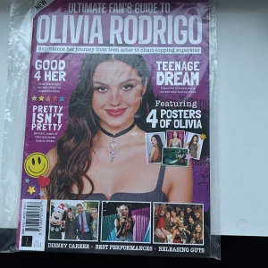 Ultimate Fan's Guide to Olivia Rodrigo - Denna tidning är en ultimat guide för fans av Olivia Rodrigo. Den har en lila omslag med bilder och artiklar om hennes karriärresa från tonårsskådespelare till superstjärna. Innehåller fyra posters av Olivia och artiklar som 'Good 4 Her' och 'Teenage Dream'. Perfekt för fans som vill veta mer om hennes liv och musik.