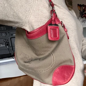Beige och röd axelväska från Prada - Säljer en snygg axelväska från Prada i beige canvas med röda skinn detaljer. Väskan har ett broderat Prada Milano-märke på framsidan och en praktisk axelrem. Perfekt för både vardag och fest! Köpt på Catawiki för 7400kr. Pris kan diskuteras vid snabb affär!🌟🥰