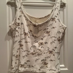 Vit blommig ribbad linnetopp - Säljer en söt vit ribbad linnetopp med blommigt mönster. Toppen har en rund halsringning och små knappar framtill. Perfekt för sommardagar eller som ett lager under en kofta. Den är i mycket bra skick och passar till allt