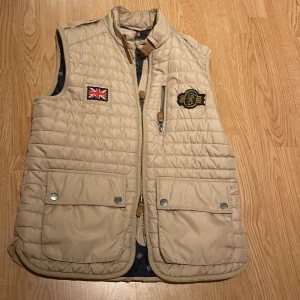 beige morris quiltad väst  -  Västen har en dragkedja och två stora fickor med knappar framtill. Perfekt för höst och vår när du vill hålla dig varm men ändå se stilren ut. Passar bra till både vardag och lite mer uppklädda tillfällen.