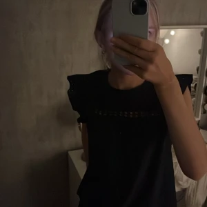 Svart blus med spetsdetaljer - Säljer en elegant svart blus med spetsdetaljer vid halsen. Blusen har korta ärmar med volang och en lös passform, perfekt för både vardag och fest. Den är i mycket bra skick och passar till många olika outfits.