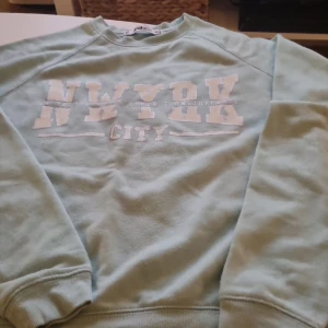 Ljusblå sweatshirt från FB Sister - Säljer en ljusblå sweatshirt från FB Sister i storlek XS. Tröjan har ett tryck med texten 'NYC' på framsidan. Den är långärmad och har ribbade muddar vid ärmslut och nederkant. Observera att det finns en liten fläck på ena ärmen. Fläcken kan man tvätta bort. Perfekt för en avslappnad stil! 💙