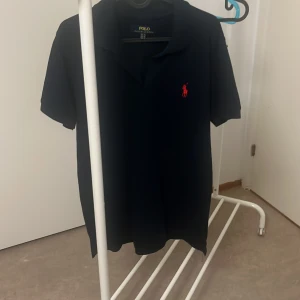 Svart kortärmad pikétröja från Polo Ralph Lauren - Säljer en svart pikétröja från Polo Ralph Lauren med en röd broderad logga på bröstet. Passar både till vardag och fest!