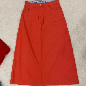 Röd kjol från U.S. Polo Assn. - Säljer en snygg orange/röd kjol från U.S. Polo Assn. Kjolen har en klassisk design med fickor både fram och bak, och en stilren knapp i midjan. Perfekt för både vardag och fest!