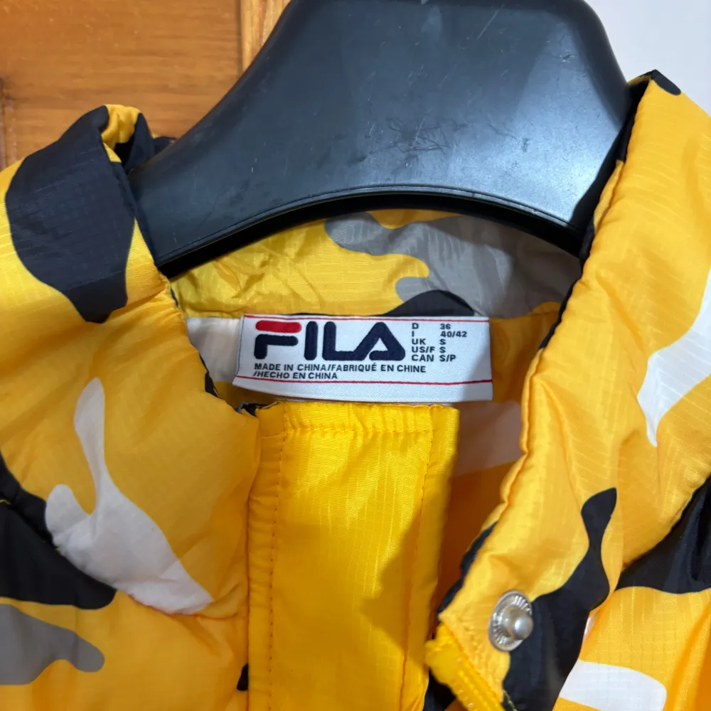 Säljer en supercool gul kamouflagemönstrad dunväst från FILA. Västen har en dragkedja framtill och en praktisk ficka med FILA-logga på bröstet. Perfekt för höst och vår när du vill sticka ut lite extra. Den är i nyskick och redo för nya äventyr!. Takit.