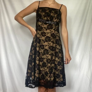 Svart spetsklänning från Reflex - Säljer en elegant svart spetsklänning från Reflex i storlek M. Jag brukar bära storlek xs eller small och den ser bra ut på mig ändå. Klänningen har ett vackert blommigt mönster och en beige underklänning som ger en fin kontrast. Den har smala axelband och en söt rosett i midjan. Perfekt för festliga tillfällen!