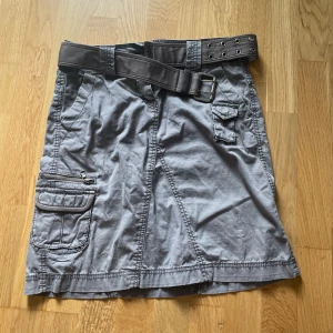 Grå kjol med bälte och fickor - Säljer en grå kjol med ett matchande bälte. Kjolen har flera praktiska fickor, inklusive en med dragkedja och en med en liten dekorativ knappdetalj.
