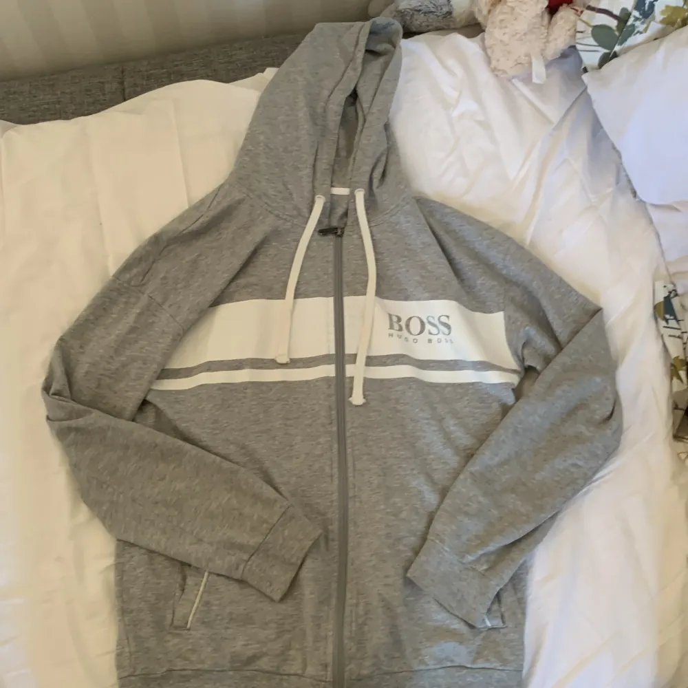 Säljer en snygg och bekväm grå hoodie från Hugo Boss. Den har en dragkedja framtill och en vit rand med loggan 'BOSS' tryckt över bröstet. Perfekt för en avslappnad stil och passar till alla tillfällen. Den är i storlek M och i topp skick pris kan diskuteras . Tröjor & Koftor.