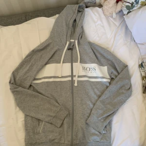 Grå hoodie från Hugo Boss - Säljer en snygg och bekväm grå hoodie från Hugo Boss. Den har en dragkedja framtill och en vit rand med loggan 'BOSS' tryckt över bröstet. Perfekt för en avslappnad stil och passar till alla tillfällen. Den är i storlek M och i topp skick pris kan diskuteras 