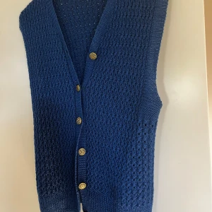 Blå stickad väst med guldknappar - Säljer en snygg blå stickad väst med fina guldknappar. Västen har en v-ringad design och ett härligt stickat mönster som ger den en unik look. Perfekt för höst och vår, och kan stylas både uppklätt och avslappnat. Passar bra för både vardag och fest!