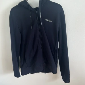 Svart hoodie från Armani Exchange - Säljer en snygg svart/marinblå hoodie från Armani Exchange. Den har en dragkedja framtill och en liten logga på bröstet. Hoodien är i storlek S och har långa ärmar. Perfekt för alla årstider! hör av er vid frågor📩