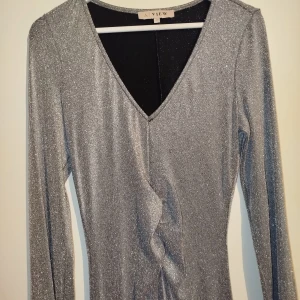 Glittrig långklänning från A View - Säljer en superfin glittrig långklänning från A View. Klänningen är i en snygg silverfärg och har en v-ringad hals. Den är långärmad och perfekt för festliga tillfällen. Materialet är syntet och den har en tight passning som framhäver figuren. Klänningen är i nyskick och redo för att användas på nästa fest eller nyårsfirande!