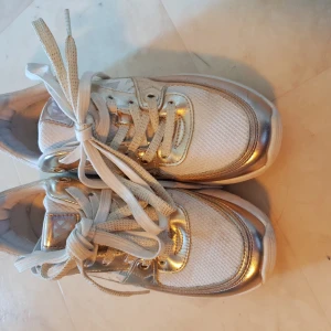 Guldiga sneakers från Michael Kors - Säljer ett par snygga sneakers från Michael Kors i vitt och guld. Skorna har en glittrig finish med snörning och en bekväm sula. Perfekta för att ge din outfit lite extra bling! Stora i storleken. 