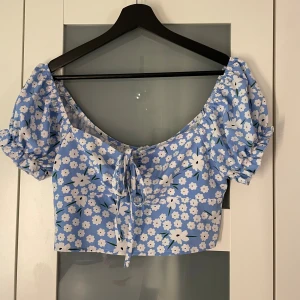 Blommig topp - Blommig topp i blått, vitt och grönt.  Kan dras åt över bysten, dragkedja i ryggen.   Sparsamt använt, i fint skick förutom små foundationfläckar på snören. Har ej försökt få bort, kanske går. Syns inte när man använder tröjan. 