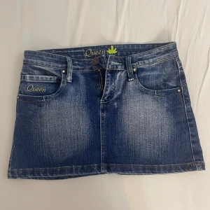 Jeanskjol från Queen - Säljer en snygg jeanskjol från Queen i storlek M (sitter på mer som ett S).Kjolen är kort och har en klassisk blå jeansfärg med slitningar för en cool look. Den har två fickor fram och två bak, med en dragkedja och knapp framtill. Det finns broderade detaljer med texten 'Queen' och en guldfärgad lövdetalj. Perfekt för en avslappnad stil!