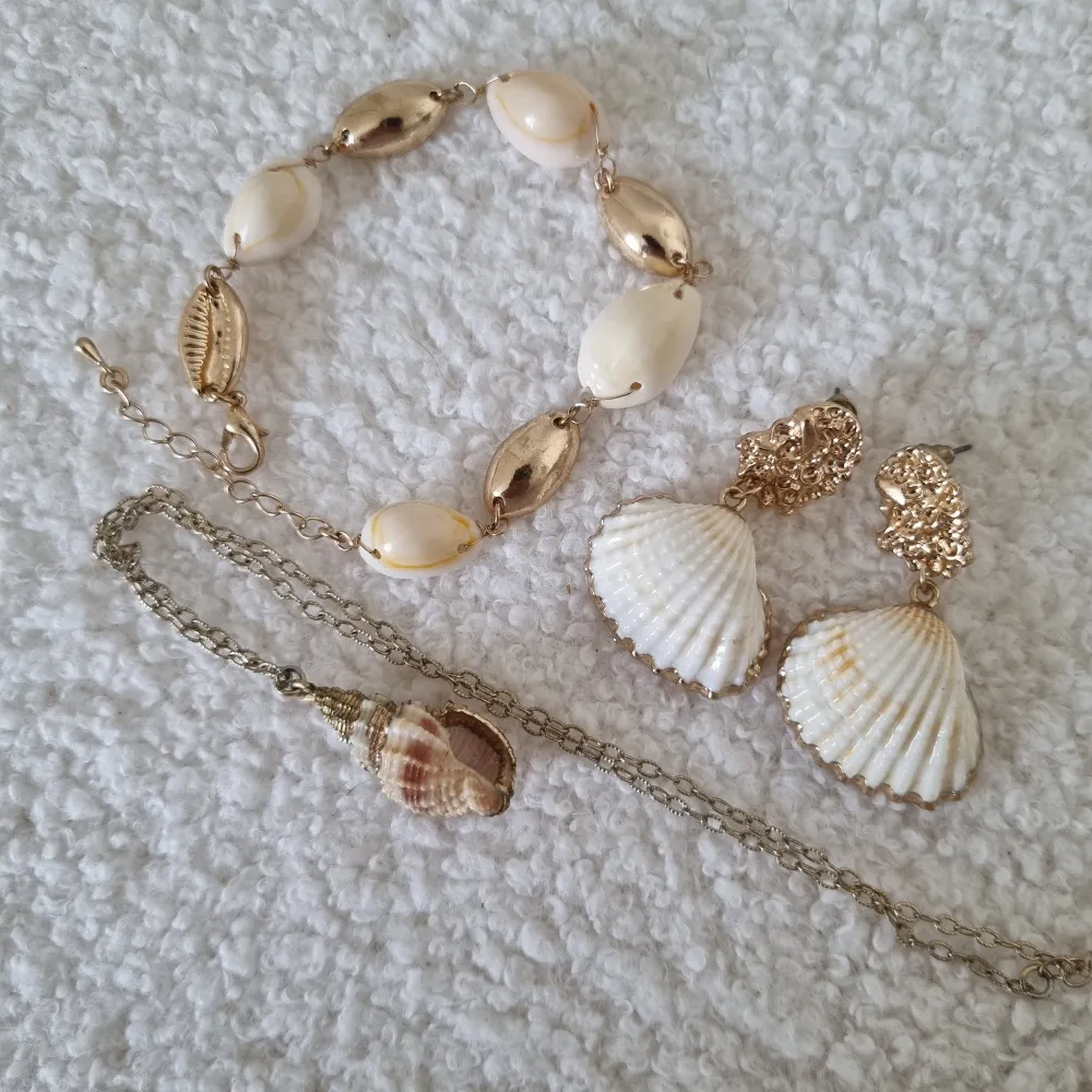 Säljer ett superfint smyckeset med snäckor! Setet innehåller ett armband/ankellänk, ett halsband och ett par örhängen. Armbandet har vita och guldiga snäckor, halsbandet har en stor snäcka som hänge och örhängena har vita snäckor med guldiga detaljer. Perfekt för sommarens alla fester och strandhäng! 🌊✨. Asusteet.