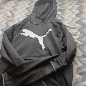 Svart hoodie från Puma - Säljer en svart hoodie från Puma i storlek M. Den är gjord av mjukt och bekvämt material, perfekt för både träning och vardagsbruk. Hoodien har en stor vit Puma-logga på bröstet och en justerbar dragsko i huvan. Den är i nyskick och har långa ärmar.