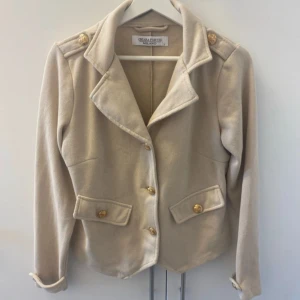 Beige kavaj från Chiara Forthi - Säljer en snygg beige kavaj från Chiara Forthi Milano. Den har en klassisk design med guldknappar och två fickor framtill. Kavajen är långärmad och har en fin passform som passar perfekt till både vardag och fest. Materialet känns mjukt och bekvämt.