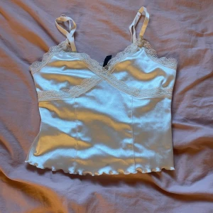 Vit satinlinne med spetsdetaljer - Säljer ett superfint vitt satinlinne med spetsdetaljer. Linnet har smala axelband och en vacker spetskant runt halsringningen. Perfekt för en somrig look eller en festkväll. Det är i nyskick och känns väldigt lyxigt på huden.