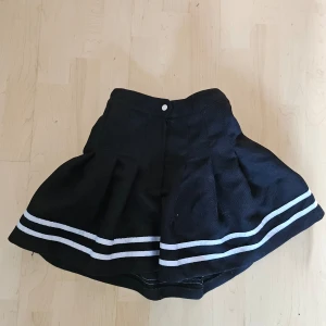 Svart kortkjol med vita ränder - Säljer en superfin svart kortkjol med två vita ränder längst ner. Kjolen har en knapp och dragkedja framtill för enkel på- och avtagning. Perfekt för både vardag och fest! 🖤