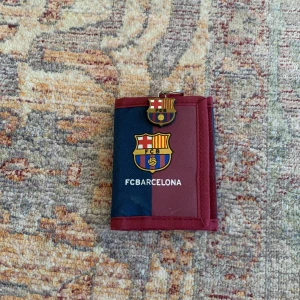 FC Barcelona plånbok - Säljer en cool FC Barcelona plånbok i blått och vinrött. Plånboken har klubbens logga på framsidan och en praktisk dragkedja inuti. Perfekt för alla Barça-fans! 💙❤️