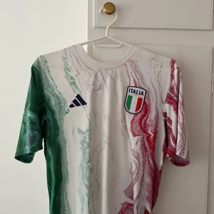 Italiensk fotbollströja från Adidas - Säljer en snygg italiensk fotbollströja från Adidas i storlek M. Tröjan har ett coolt marmorliknande mönster i grönt, vitt och rött, vilket representerar Italiens flagga. Den är i nyskick och har korta ärmar. Perfekt för fotbollsfans eller som en stilren sporttröja!