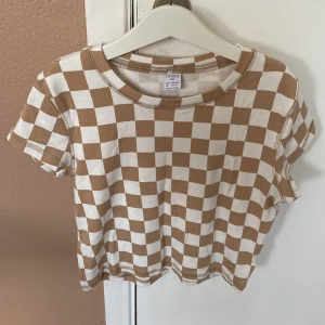 Brun och vit rutig t-shirt från Lindex - Säljer en supercool brun och vit rutig t-shirt från Lindex. Den är i storlek 158/164 och passar perfekt för någon som är 12-14 år. T-shirten är kortärmad och croppad, vilket gör den perfekt för sommaren. Den är i nyskick och gjord av bomull, så den är både bekväm och snygg. Perfekt för en avslappnad och trendig look! köparen betalar för frakt 