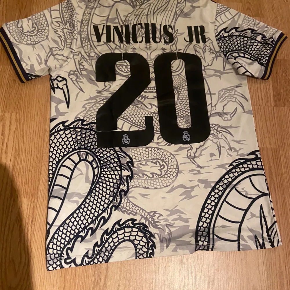 Säljer en unik Real Madrid fotbollströja från Adidas med ett coolt drakmönster. Tröjan är kortärmad och har en stilren design med mörk blå krage och detaljer. Perfekt för fans av Vinicius Jr, då hans namn och nummer 20 pryder baksidan. Passar perfekt för både match och vardag! Pris kan diskuteras. . T-shirts.