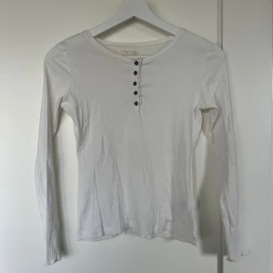 Vit ribbad långärmad topp - Säljer en stilren vit ribbad topp med långa ärmar och knappar framtill. Perfekt för både vardag och festliga tillfällen. Toppen är i bra skick och har en skön passform.