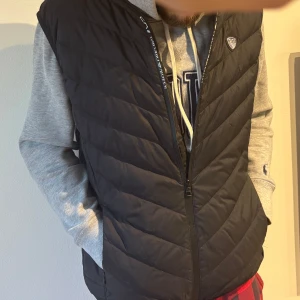 Svart quiltad väst från Emporio Armani - Säljer en snygg svart quiltad väst från Emporio Armani. Den har en dragkedja framtill och ett stilrent märke på bröstet. Perfekt för lager-på-lager under höst och vår. Passar både till vardags och lite finare tillfällen. Jag säljer den då den är lite stor för mig.