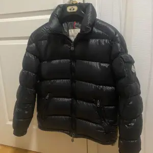 Säljer en snygg svart pufferjacka från Moncler i mycket bra skick. Storlek M passar S, Jackan är perfekt för vintern med sitt quiltade mönster och långa ärmar. Den har en dragkedja framtill och praktiska fickor med dragkedjor. Moncler-loggan är broderad på ärmen. Perfekt för att hålla sig varm och stilren i kylan! Jackan har en liten skada vid vänster ficka som syns på bilden!