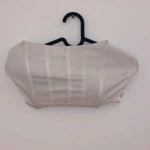 Vit korsettopp - Säljer en stilren vit korsettopp i sidenmaterial. Toppen är axelbandslös och har en tight passform som ger en elegant look. Perfekt för festliga tillfällen eller en utekväll. Den är i mycket bra skick och redo att bäras av någon som vill sticka ut.