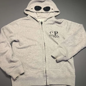 Grå hoodie från C.P. Company - Säljer en grå hoodie från C.P. Company i bra skick. Den har en cool design med inbyggda glasögon i huvan och dragkedja framtill. Perfekt för höst och vår när du vill ha något bekvämt och stiligt. Storleken är 10, vilket motsvarar en medium i barnstorlek. Passar perfekt för både chill och casual outfits! Nypris 2700kr ~ mitt pris 1800🤩🤩