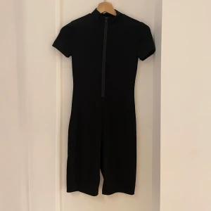 Svart kortärmad jumpsuit - Snygg och stilren svart jumpsuit med korta ärmar och dragkedja framtill. Perfekt för en minimalistisk look, passar både till vardags och festligare tillfällen. Materialet ser ut att vara stretchigt och bekvämt.