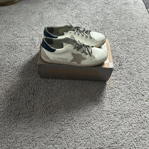 Golden Goose sneakers i vitt och grått - Säljer ett par Golden Goose sneakers i vitt med grå detaljer och en stjärna på sidan. Skorna har en cool, sliten look och blå häl. Perfekta för en avslappnad stil och passar till både jeans och shorts. Kommer i originalkartong.