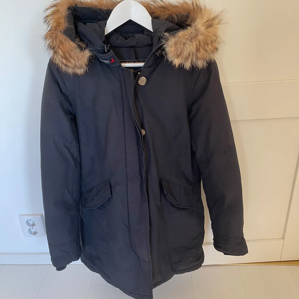 Säljer en blå dunjacka från Woolrich i mycket bra skick. Jackan har en mysig pälshuva, perfekt för kalla vinterdagar. Den har både dragkedja och knappar. I fint skick med tecken från användning. Artic parka Woolrich. . Jackor.