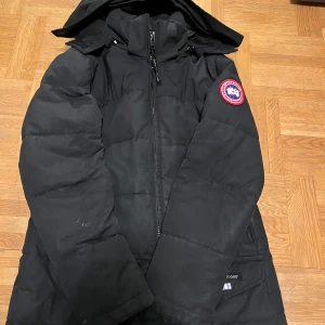 Svart dunjacka från Canada Goose - Säljer en svart dunjacka från Canada Goose i storlek XS. Jackan är perfekt för kalla vinterdagar med sin tjocka vaddering och huva. Den har en dragkedja framtill och en ikonisk logga på ärmen. Perfekt för att hålla sig varm och stilren under vintern! ❄️