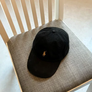 Svart keps från Polo Ralph Lauren - Snygg svart keps från Polo Ralph Lauren med den klassiska broderade loggan i guld framtill. Kepsen har en justerbar rem baktill för perfekt passform. Perfekt för både vardag och sportiga aktiviteter. En tidlös accessoar som passar till allt!