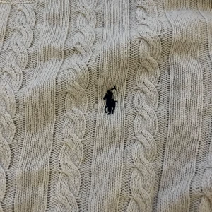 Beige stickad tröja från Ralph Lauren - Säljer en snygg beige stickad tröja från Ralph Lauren med klassiskt flätmönster. Tröjan har långa ärmar och en broderad logga på bröstet. Perfekt för kyliga dagar och ger en stilren look. Passar både till vardags och lite finare tillfällen. Tröjan har ett litet hål på vänstra armen kom dm för bild. Tröjan är xl men passar L